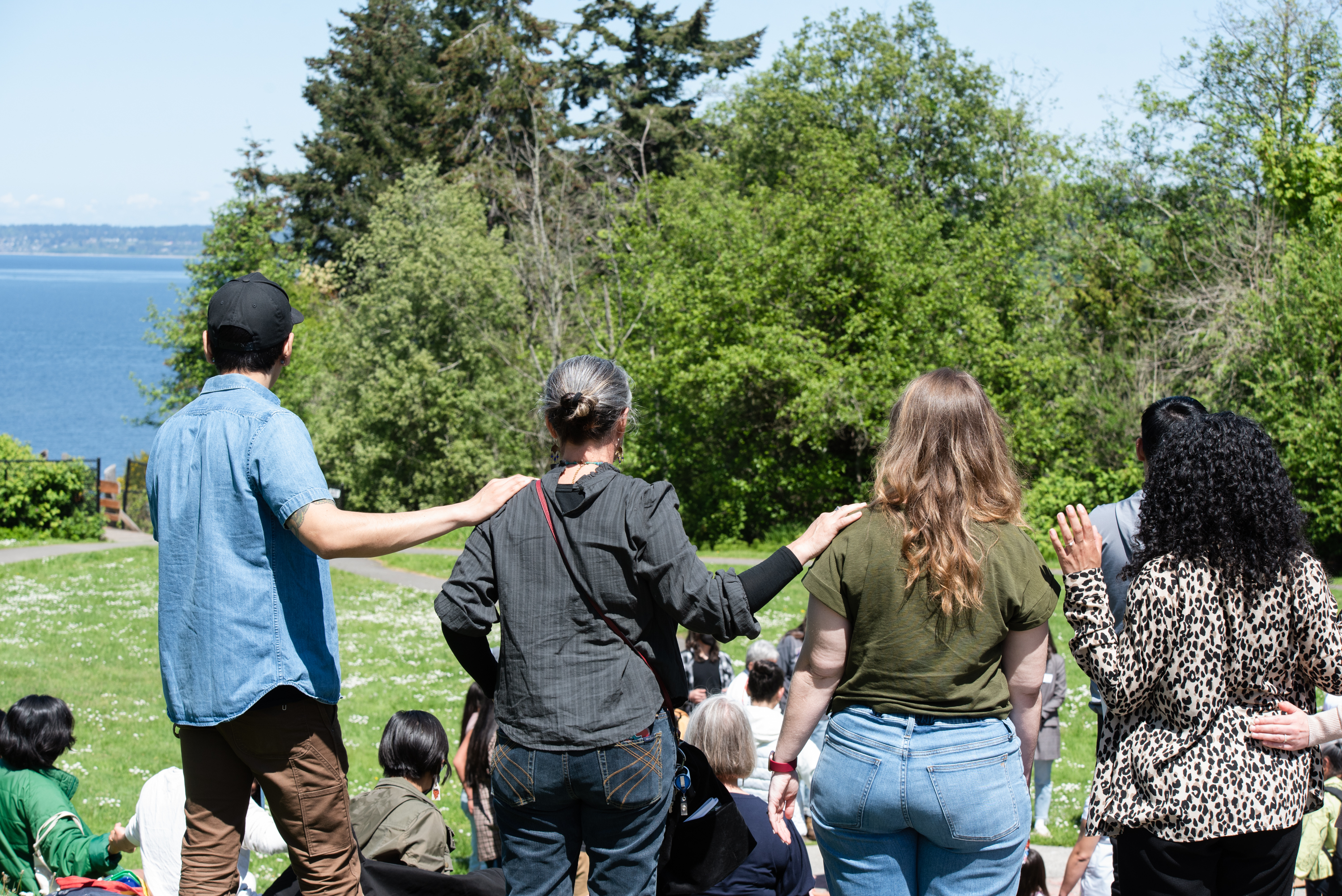 Photo of a group of people outside against a background of a blue sky and large green trees and a green lawn, with in the foreground 4 people standing in a line with their backs to the camera with their hands on each others shoulders