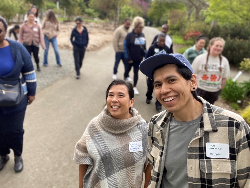 Two smiling people in focus, with other walking behind them out of focus, in an outdoor setting. 