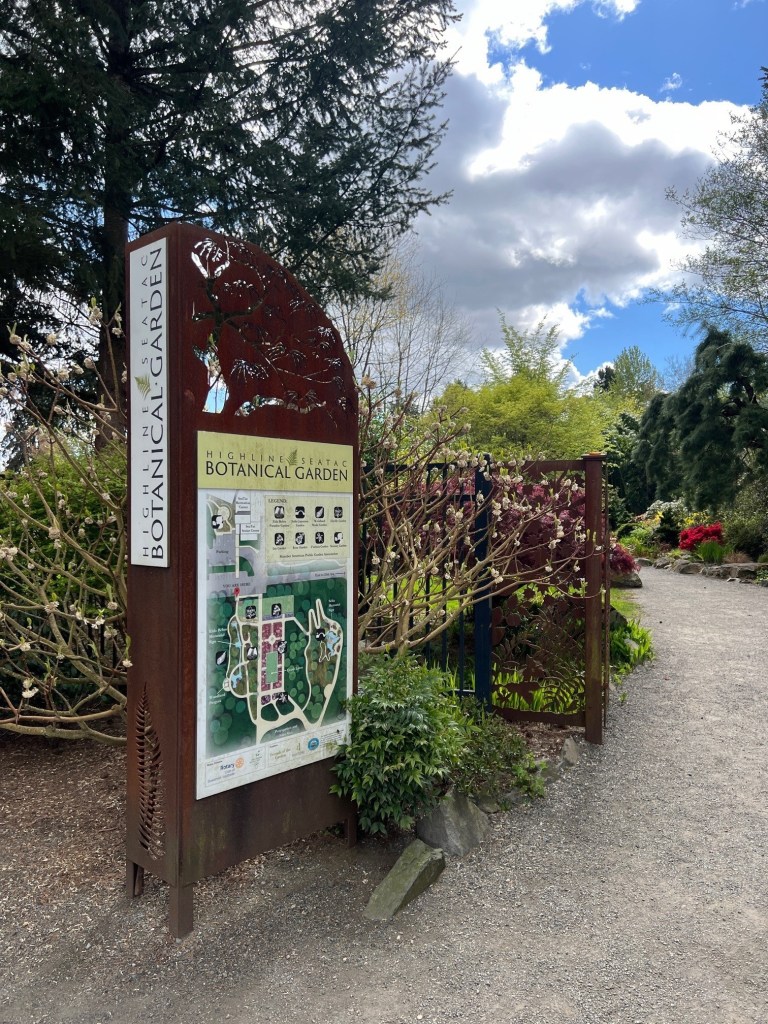 A sign reading "Highline SeaTac Botanical Garden" with a map to the garden is posted near a tree and fence in a garden area.
