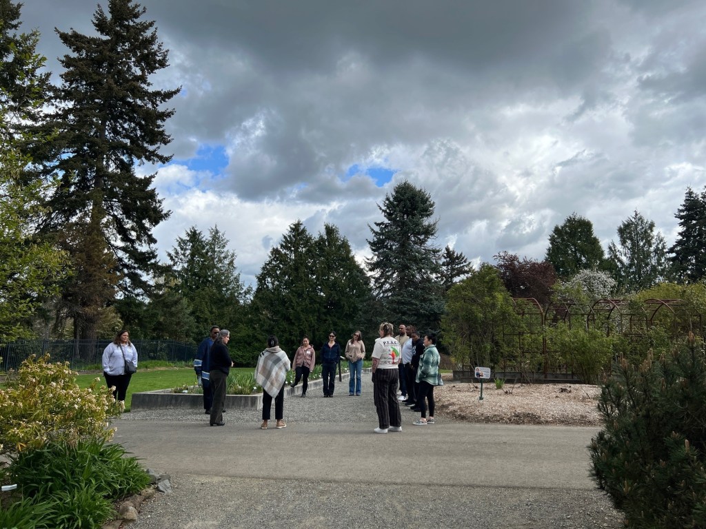 A group of people stands in a circle in an outdoor garden area on a partly cloudy day. There are tall trees behind them.
