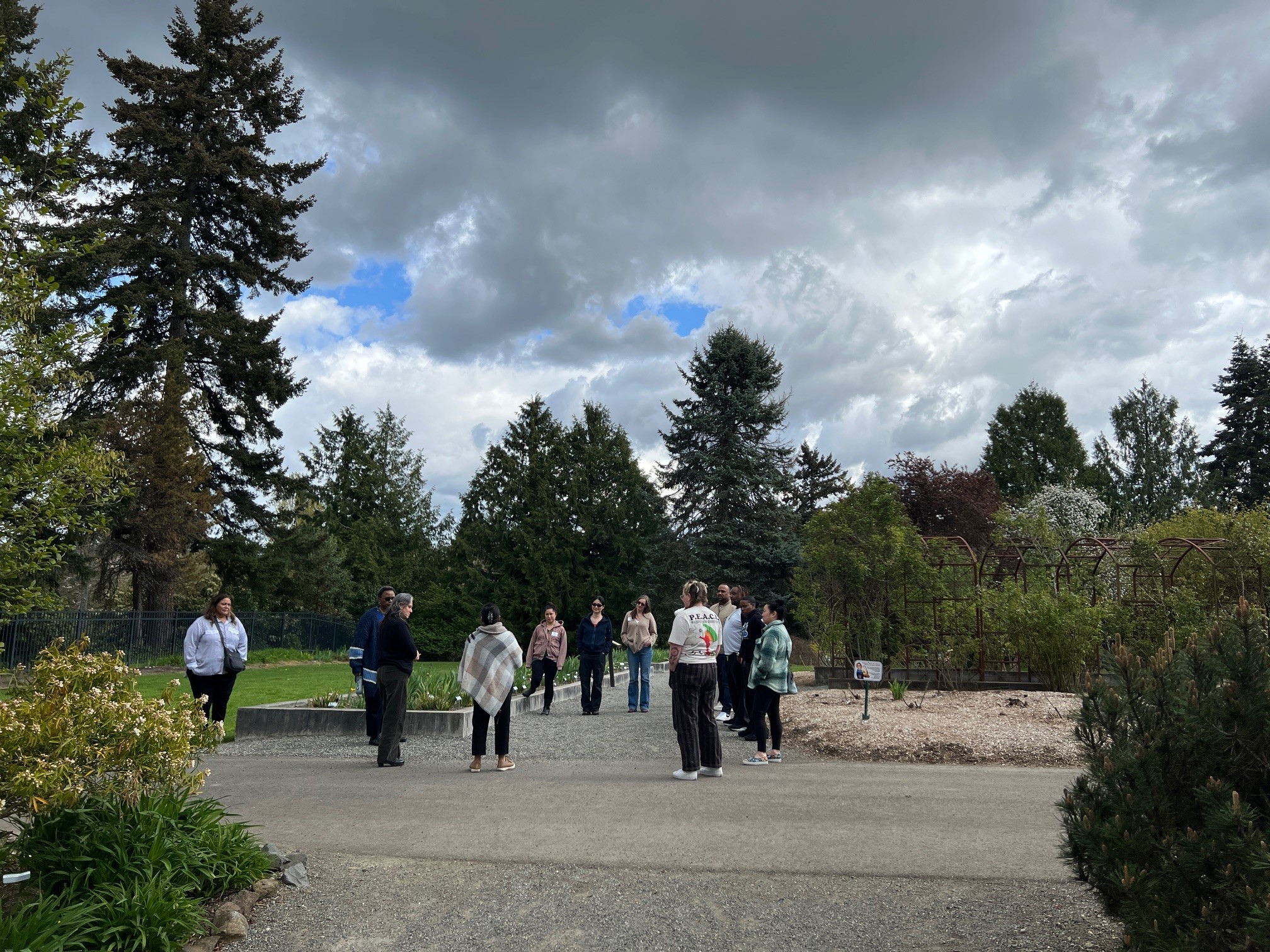 A group of people standing in a circle in an outdoor garden setting on a partly cloudy day. There are tall trees in the background.