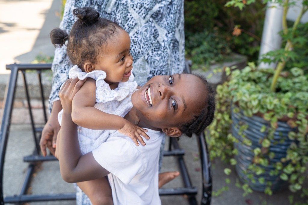 A young black child looking up and smiling at the camera, while holding black baby sibling in arms.