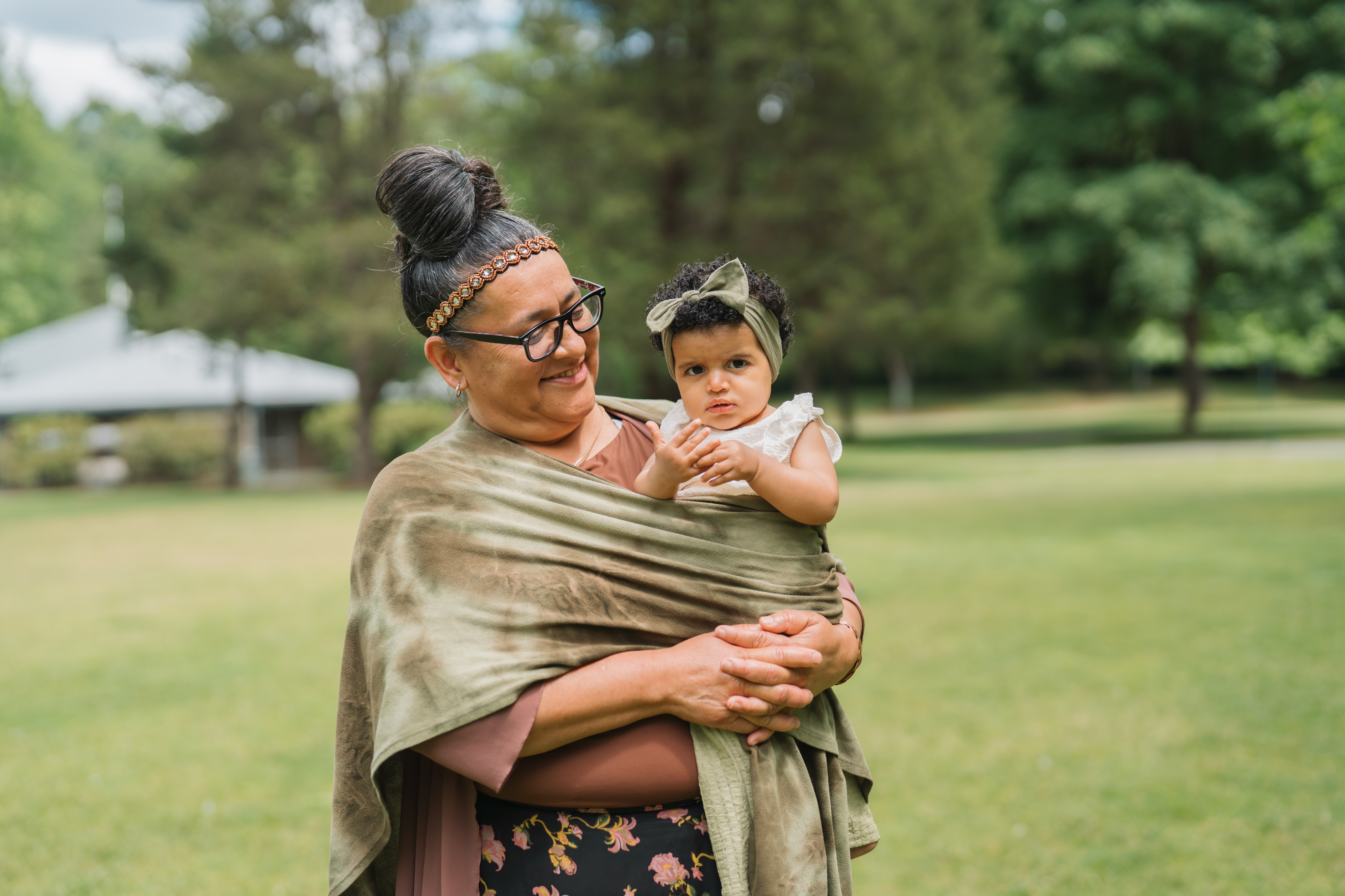 A caregiver smiling at and holding a young child in arms, wrapped in a shawl. The young child is looking at the camera.