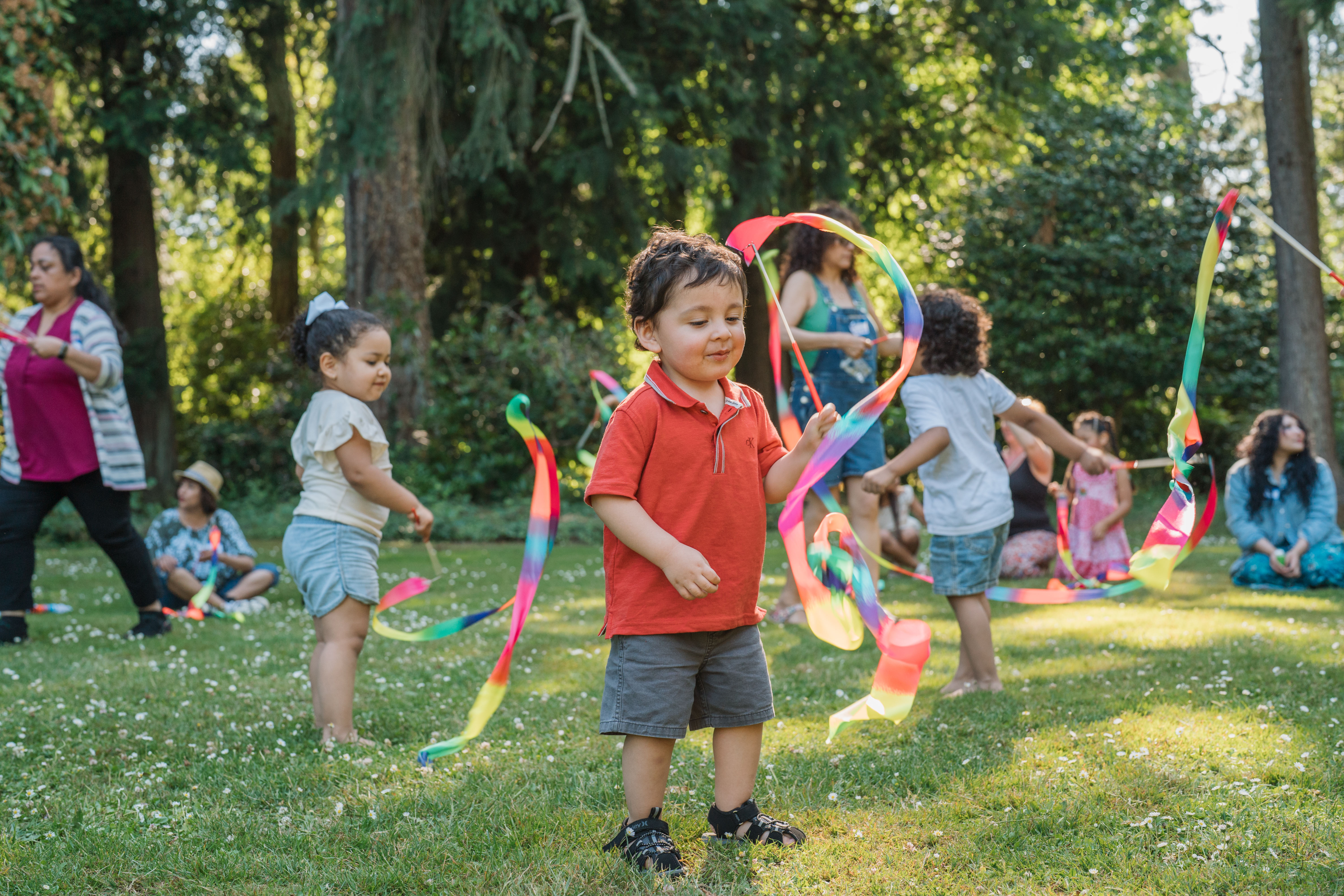 A young child twirling a ribbon wand outside. More children are playing in the background.