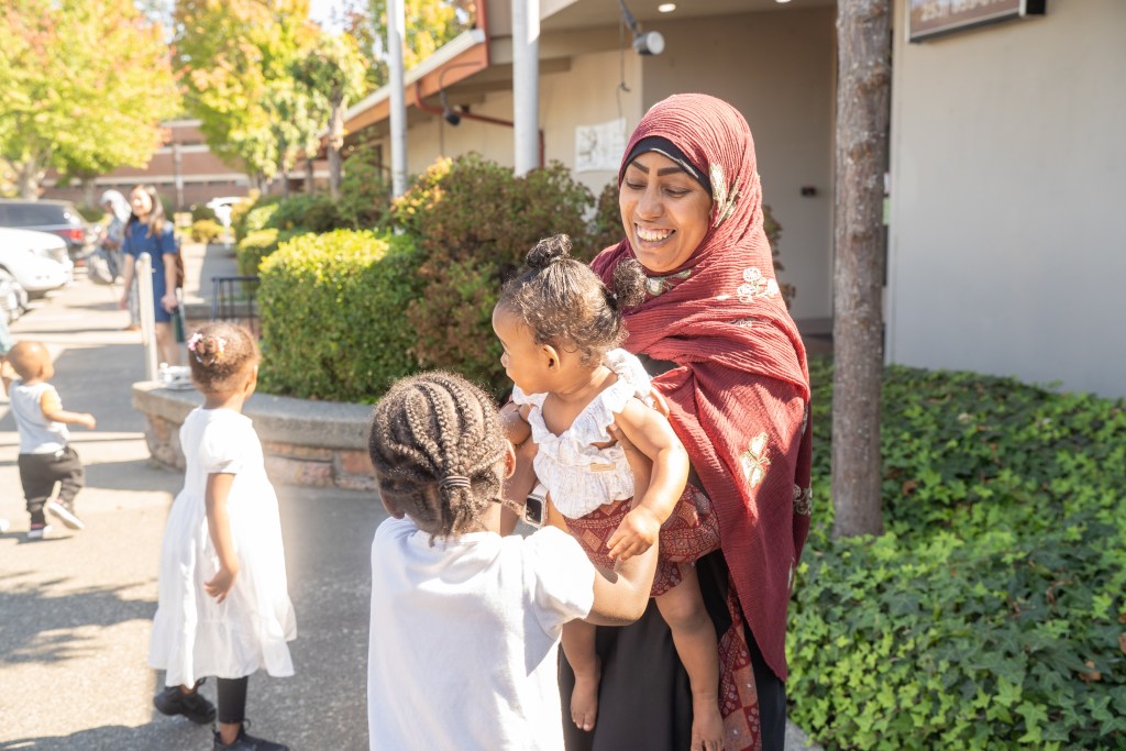 Caregiver smiling and holding young child. Children standing around outside.