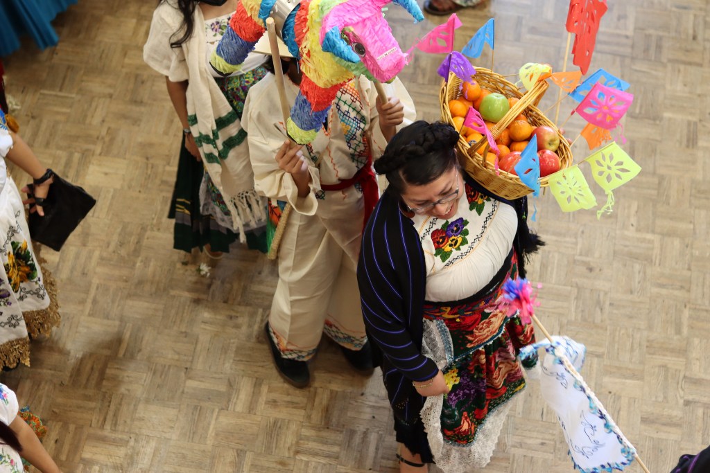 An overhead view of a an celebratory moment -- multiple people in a line with a fruit basket, a piñata, and culturally significant clothing. This photo was captured at the Resilience of Indigenous Communities event. 