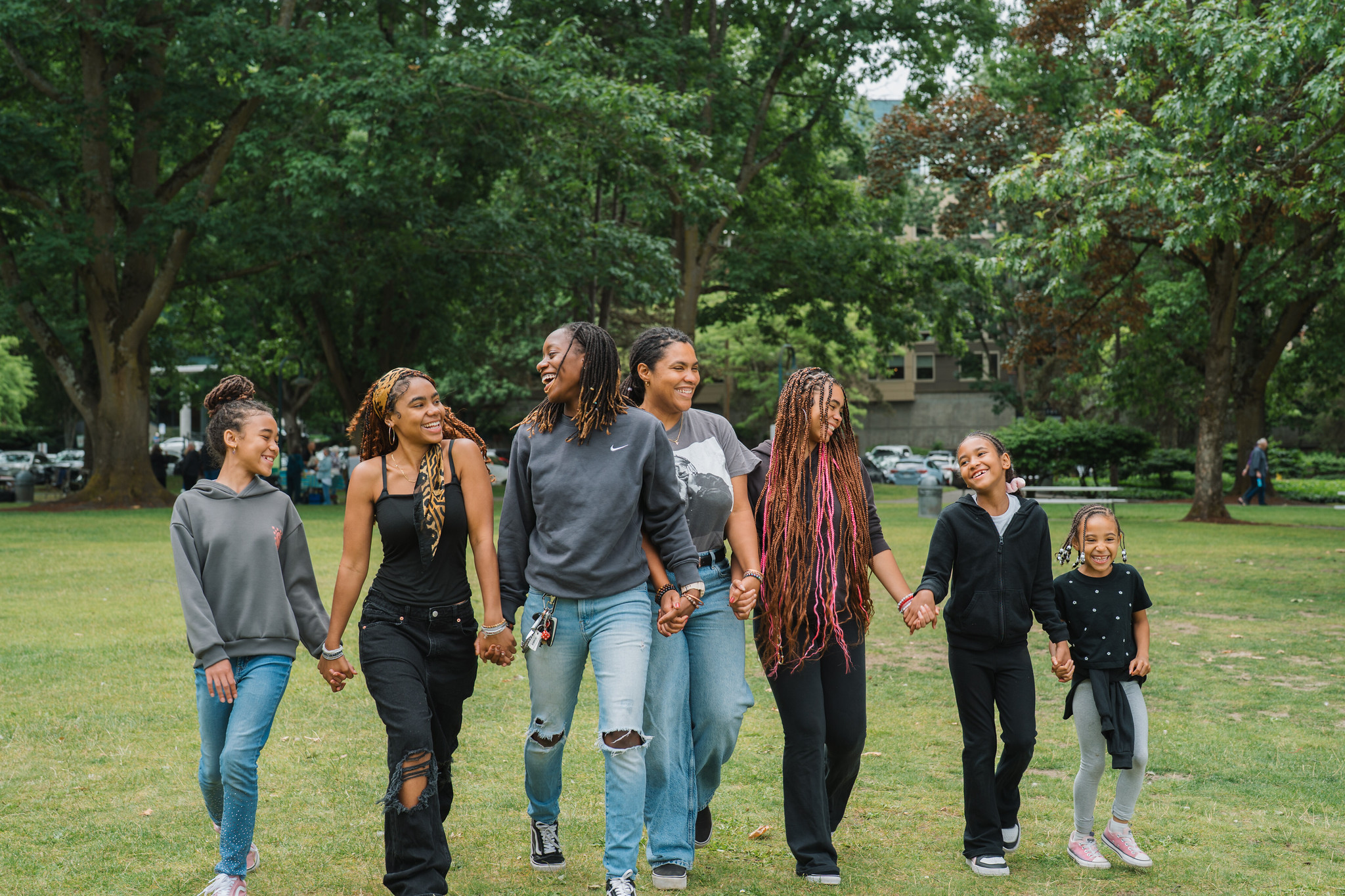 A group of young Black women and girls, smiling broadly, holding hands, and walking through a green area.